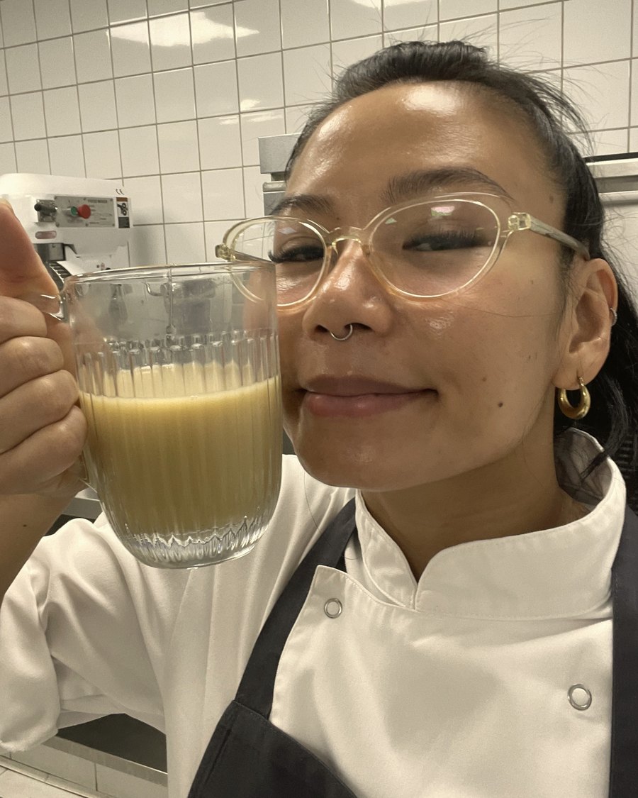 Rein Estebal in chef’s whites and a navy apron in a tiled professional kitchen, holding up a glass jug of custard.